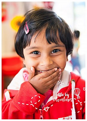 A little girl giggles behind her hand at something funny the Little Big Adventures photographer has said at her childcare